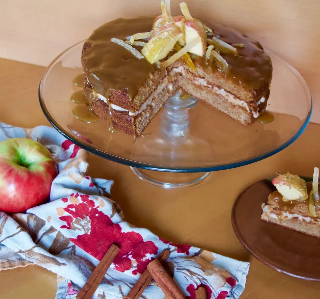 The cake on a clear glass cake stand with a two slices cut. There is a napkin with an apple and some cinnamon sticks in the foreground. One slice is on a plate to the side.