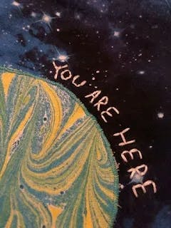 A close up of YOU ARE HERE embroidered over the Earth on a quilt of the solar system