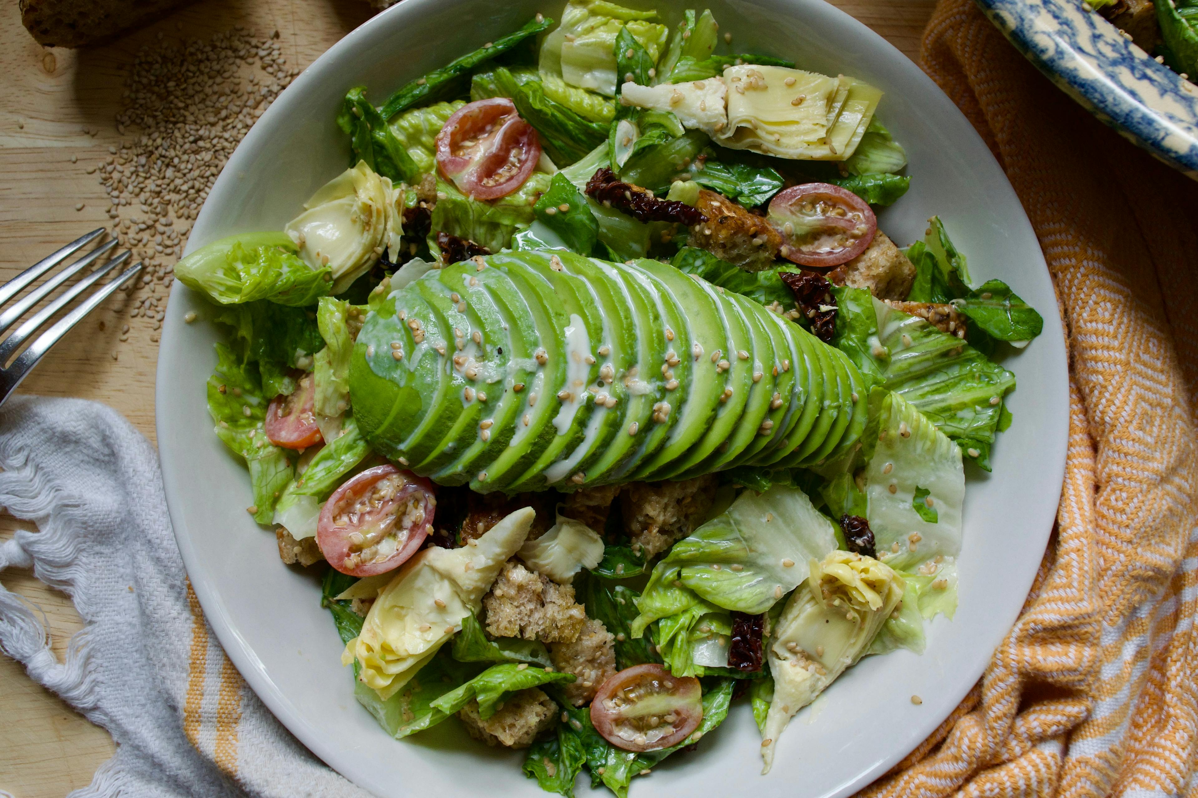 A bowl of salad with tomatoes, croutons, and artichoke hearts, with a sliced half of avocado on top.