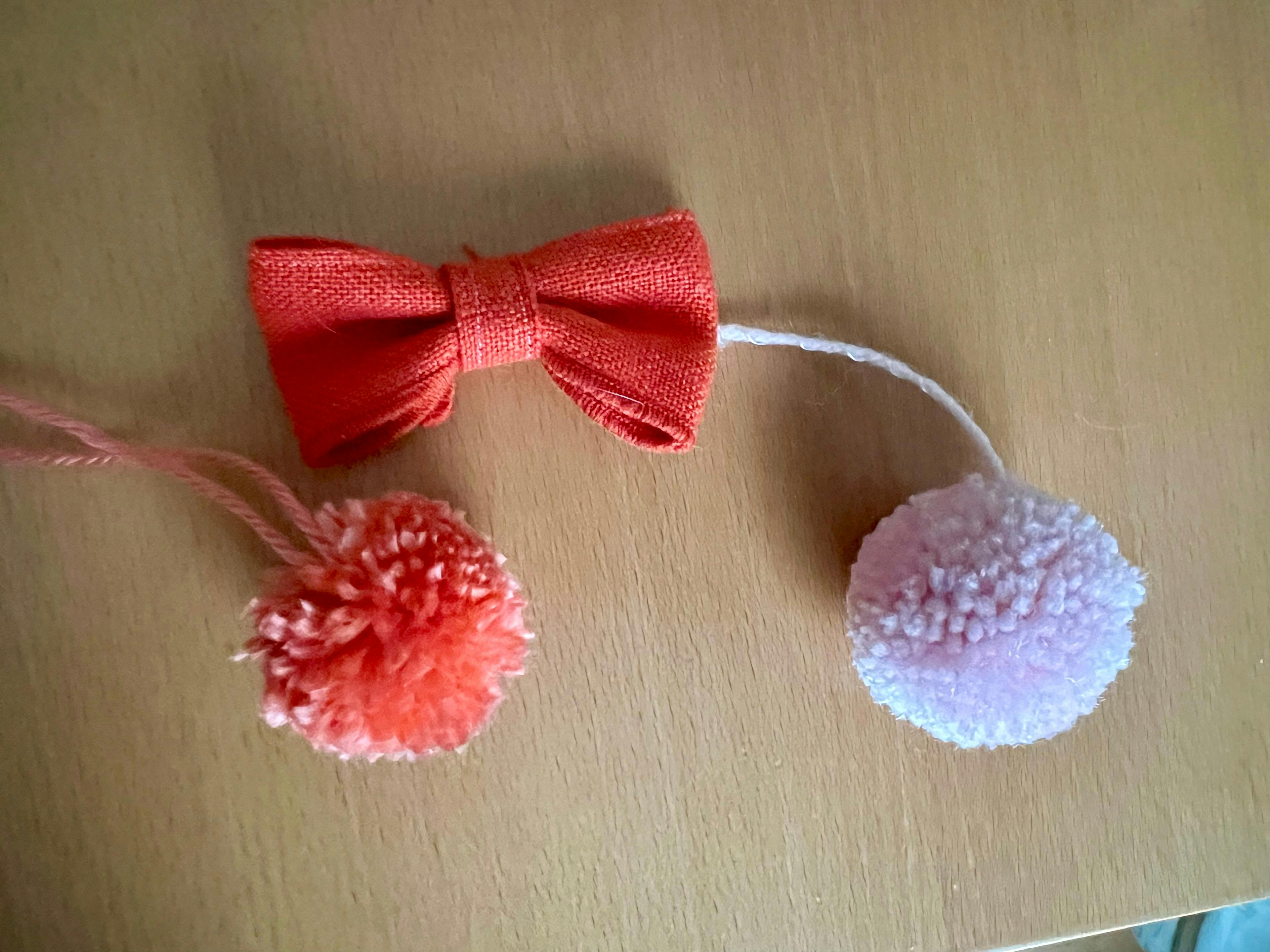 An orange fabric bow, and two pom poms made of yarn - one orange and one light pink