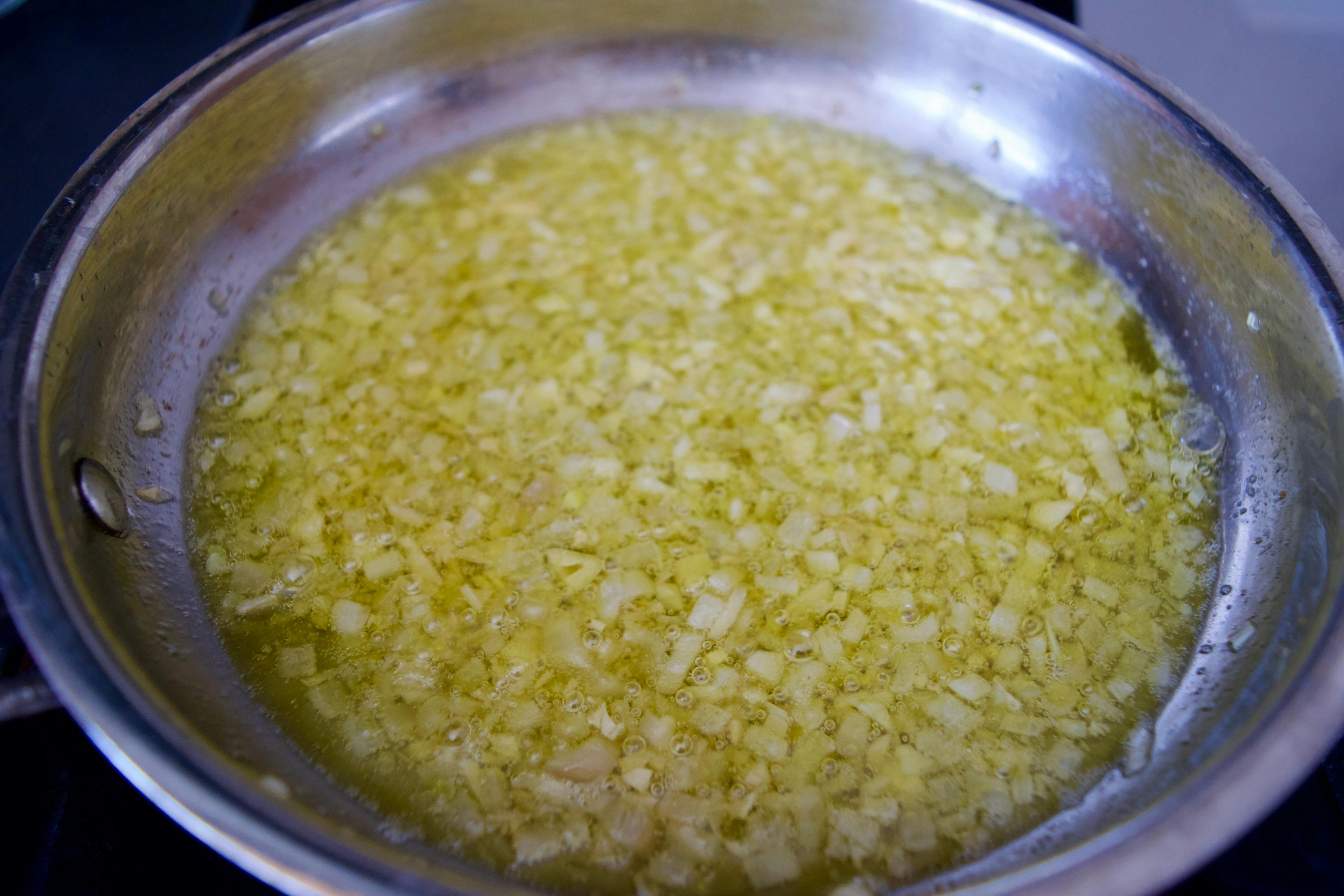 A pan of oil on a stove with minced garlic and shallots poaching. Little bubbles are visible.