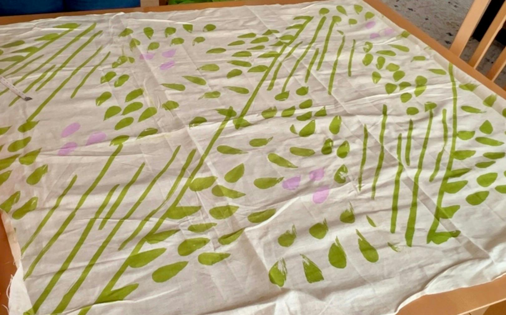 A piece of cream colored fabric with abstract brush stoke leaves and stems in green and purple is laid across a table.
