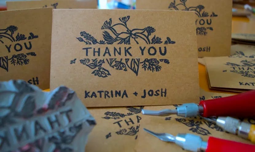 Still life of multiple brown thank you cards with black ink that says "THANK YOU" with a floral pattern behind and "KATRINA + JOSH" at the bottom of the card, thank you stamp, and carving knives.