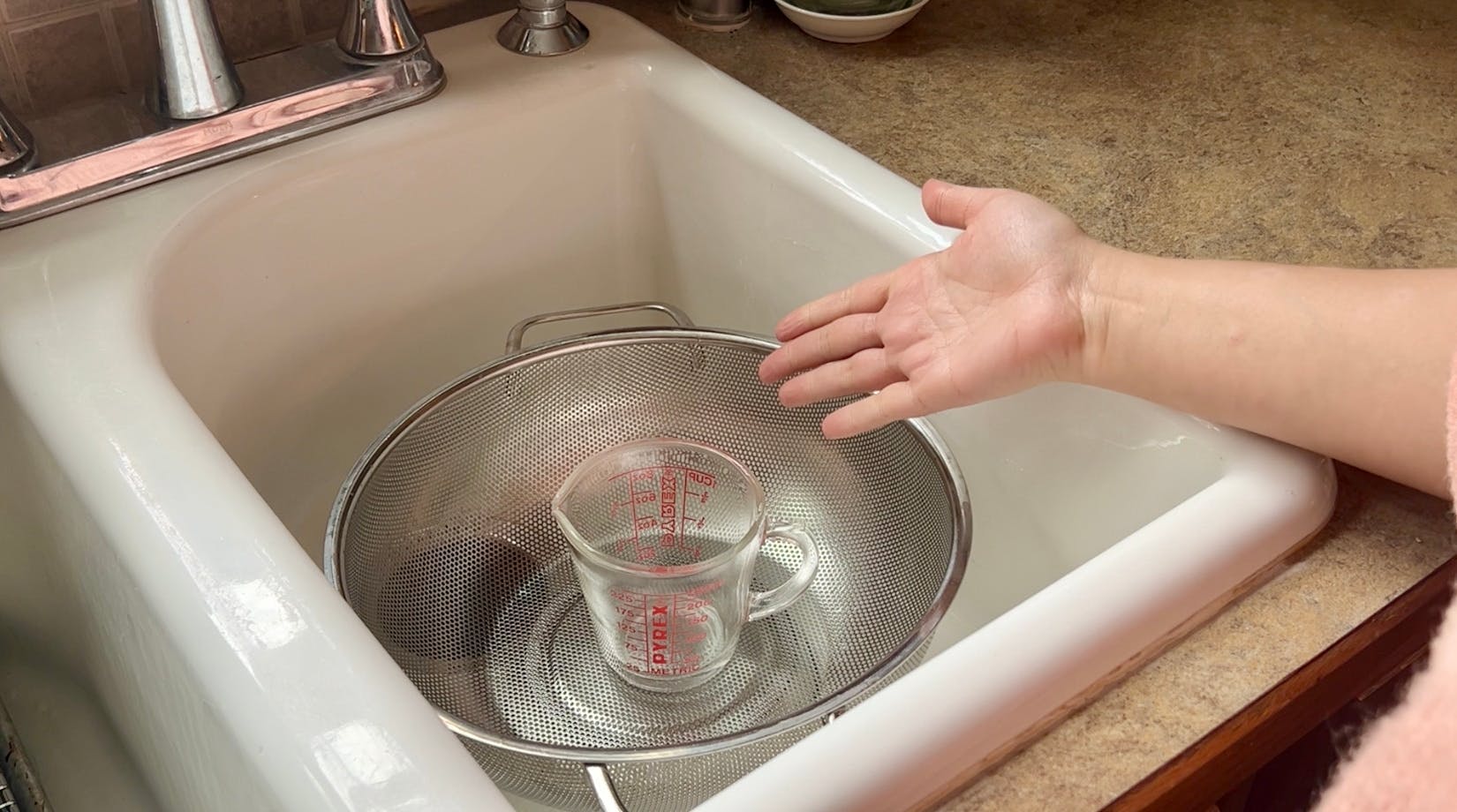 A glass measuring cup in a colander in a sink.