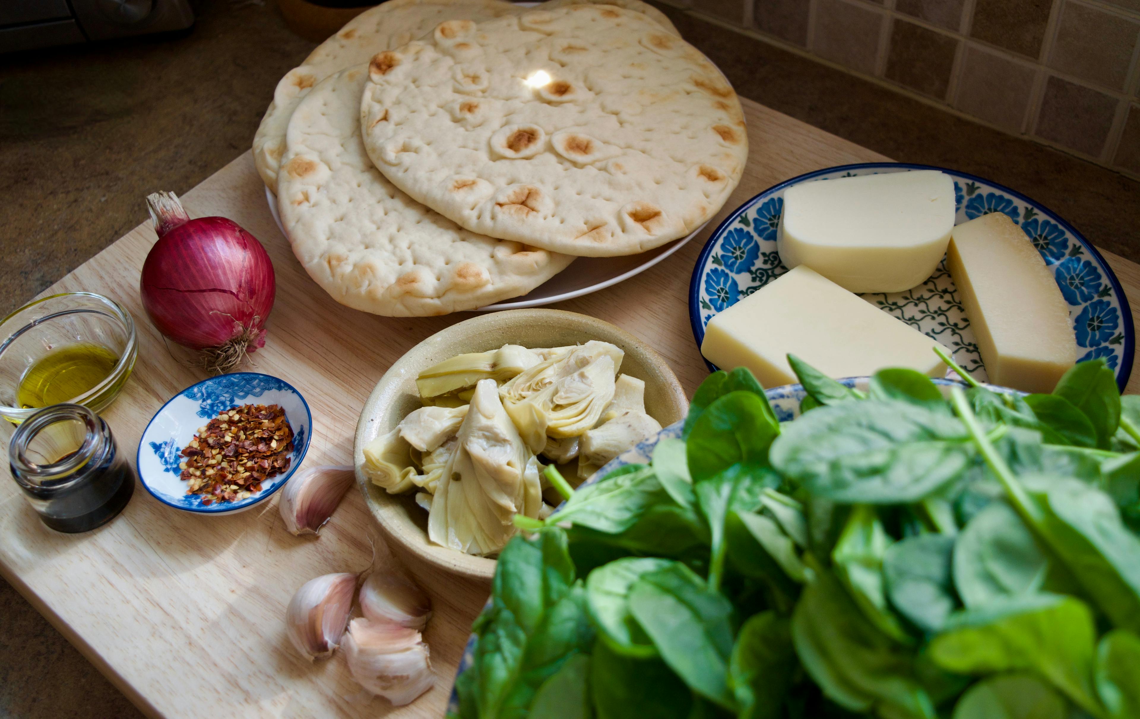 Ingredients portioned out - spinach, cheeses, artichoke hearts, garlic cloves, red onion, naan, red pepper flakes, balsamic vinegar, olive oil