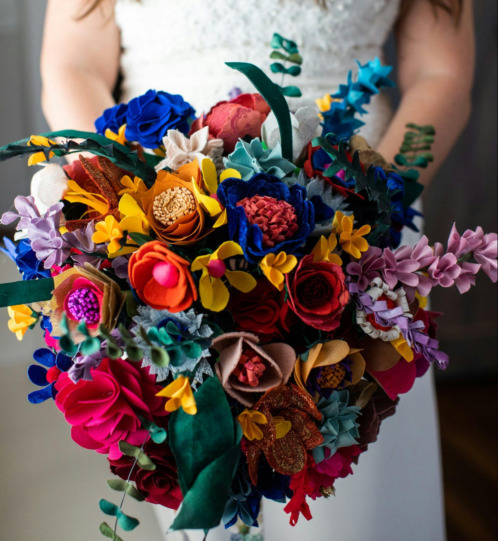 A bride in a white dress holding her bouquet. The bouquet is made of fabric flowers in various jewel tones and is quite large.