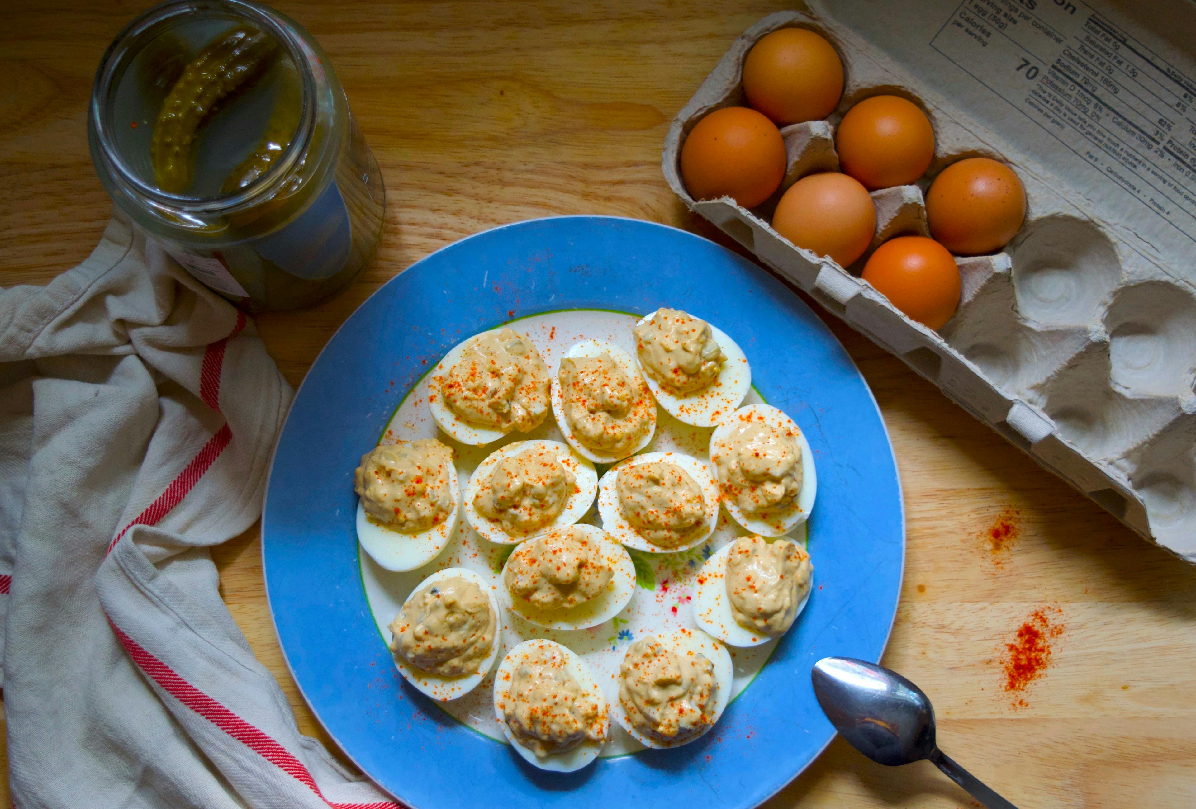 Still life with deviled eggs on a blue plate, carton of eggs to the side, jar of pickles, kitchen towel, and a spoon on the plate.