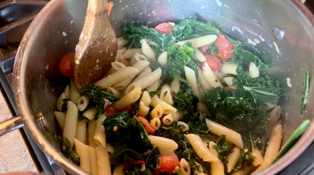 Stirring a pot of pasta, kale, and cherry tomatoes, breaking apart one of the tomatoes with the back of a wooden spoon.
