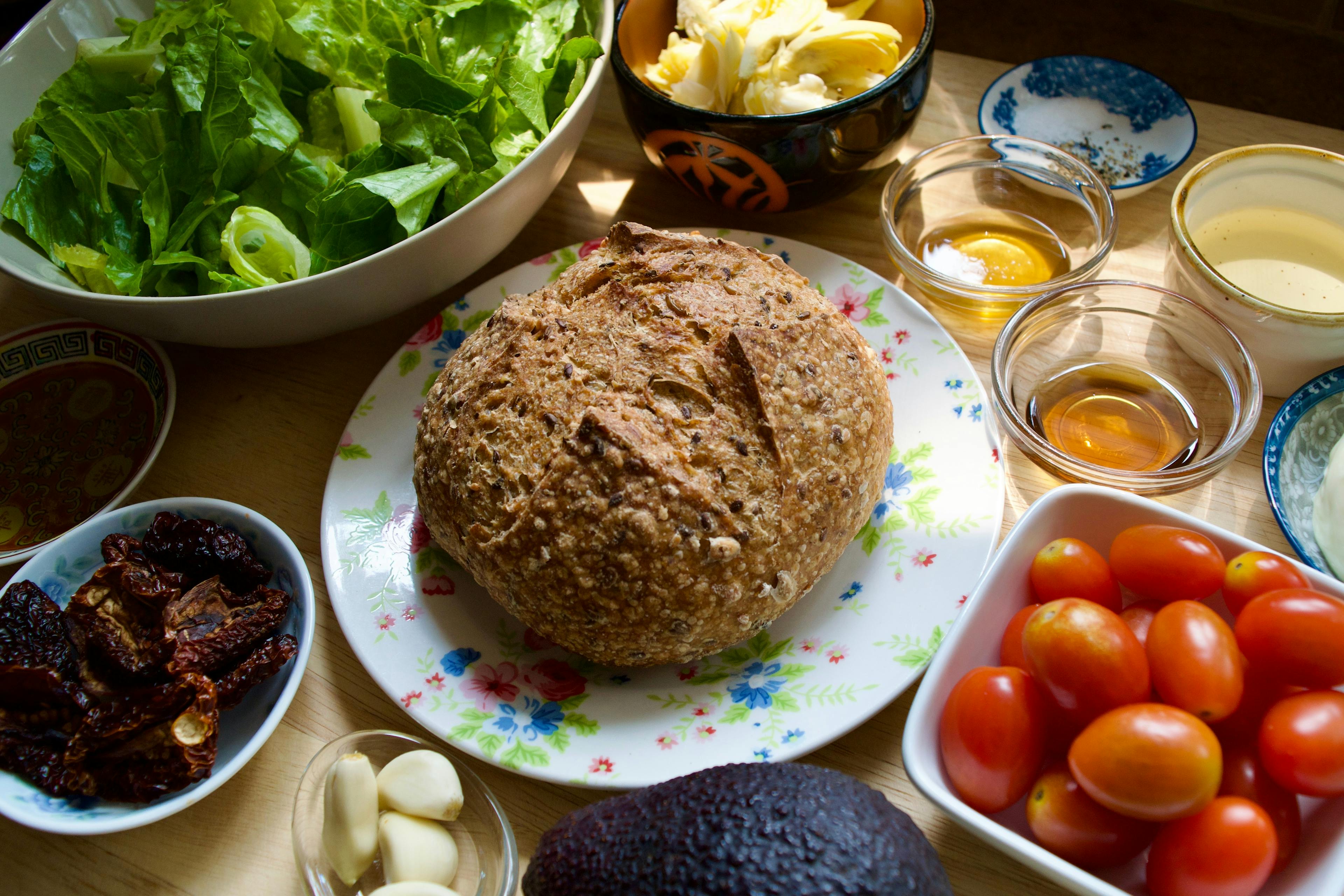 Ingredients in bowls - romaine, cherry tomatoes, garlic cloves, artichoke heart, sesame oil, sun dried tomatoes, and there is a small loaf of sourdough at the center.