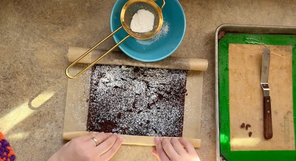 Two hands rolling a thin chocolate cake into a log.