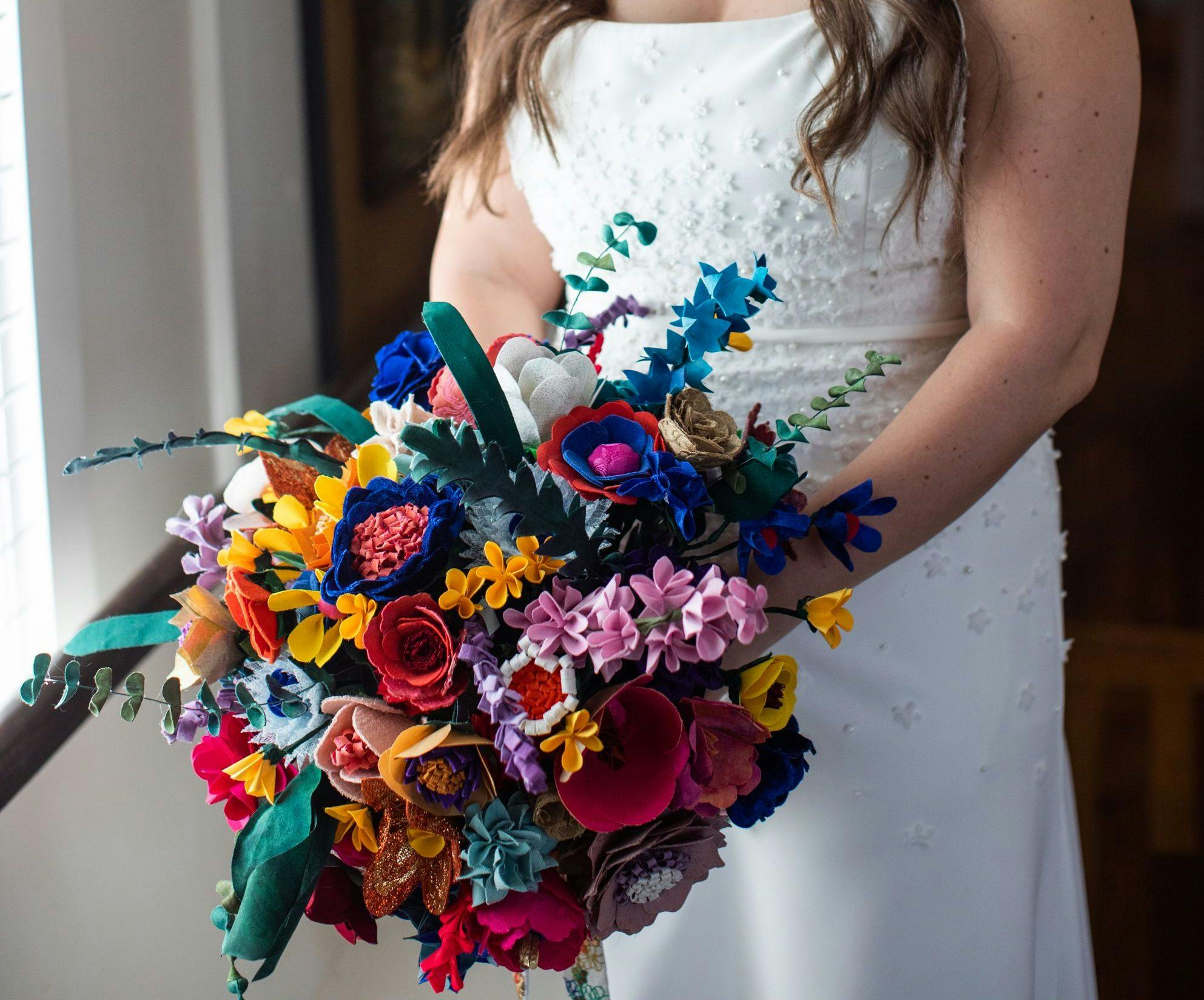 A bride with long brown hair looks down lovingly at her bouquet, which is large and made of fabric flowers in a variety of shapes and colors.