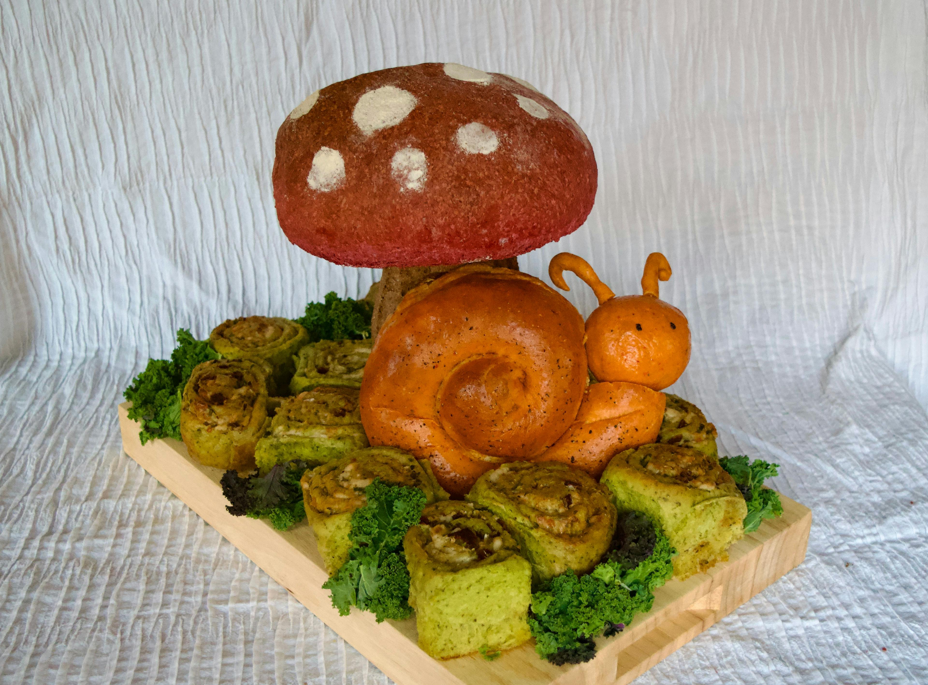 A bread sculpture on a cutting board with an orange bread snail and a red bread mushroom.