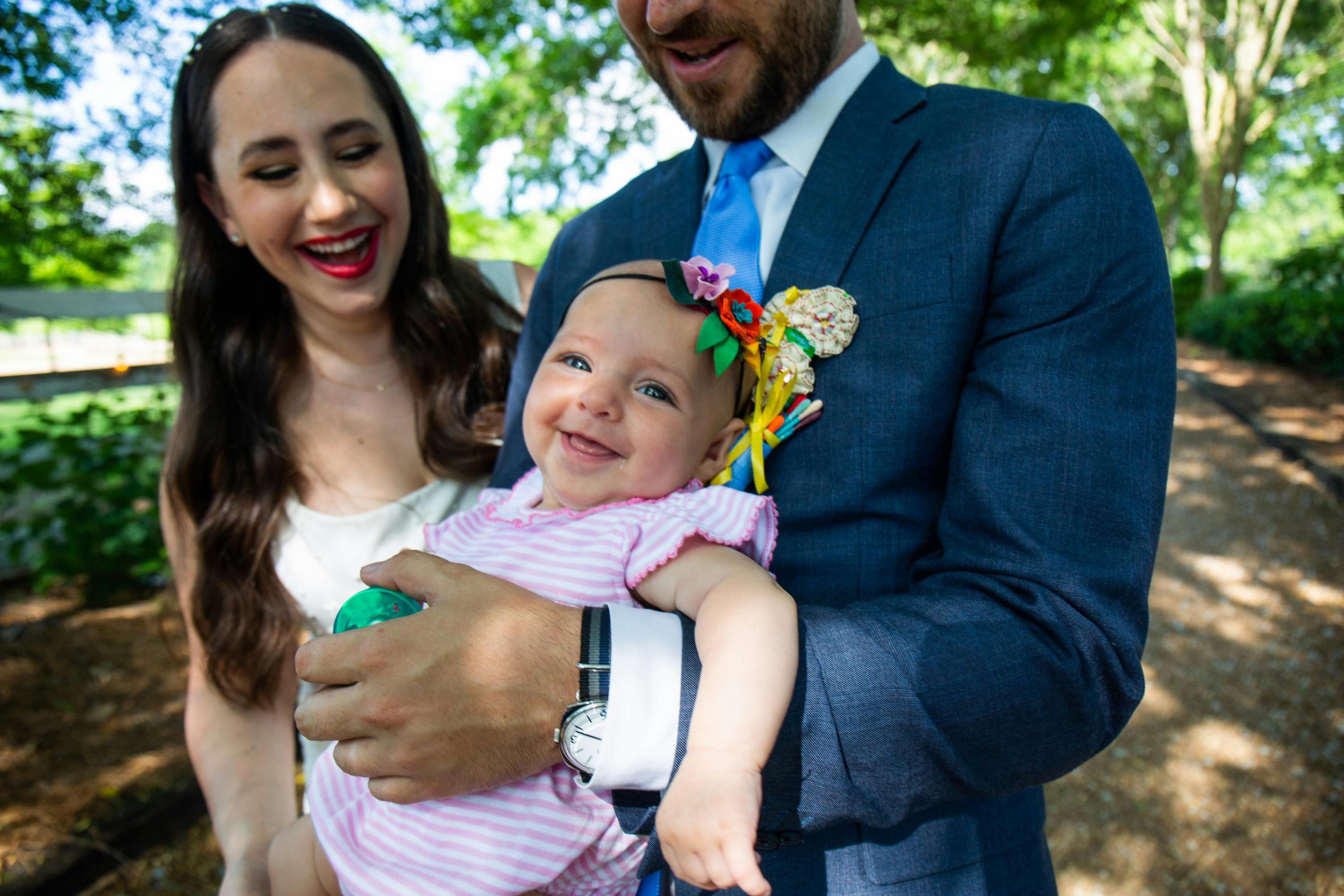 Bride Lauren in white dress, groom Jake in blue suit, and 3 month old daughter Rhea in striped pink and white onesie on the wedding day. All three are smiling, and Rhea has on a headband with three more fabric flowers.