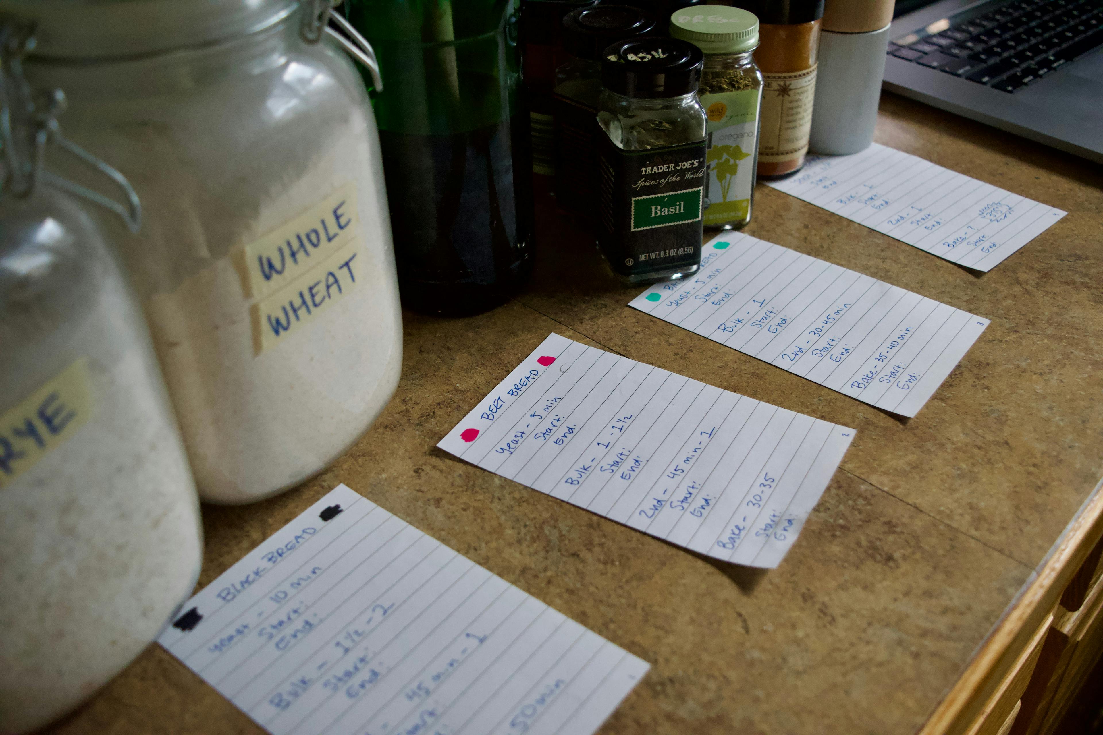 Four color coded notes with rise and bake times written on them, plus flour and bottles of dried herbs on the background