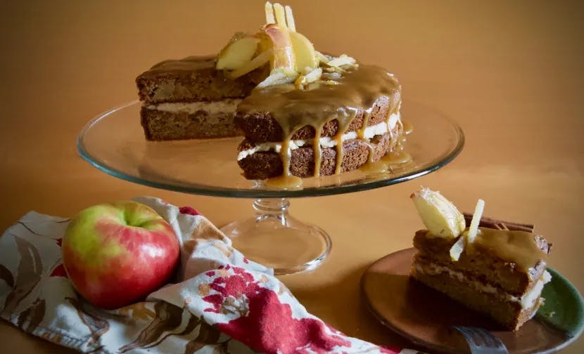 A brown spiced cake with white frosting and toffee sauce drizzled on top on a clear glass cake stand with a two slices cut. There is a napkin with an apple and some cinnamon sticks in the foreground. One slice is on a plate to the side.