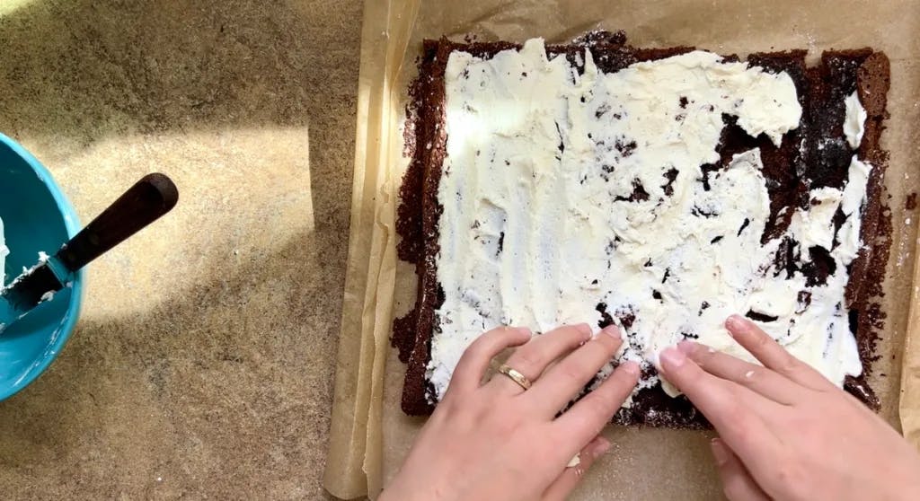 Two hands spreading white frosting onto a thin chocolate cake.