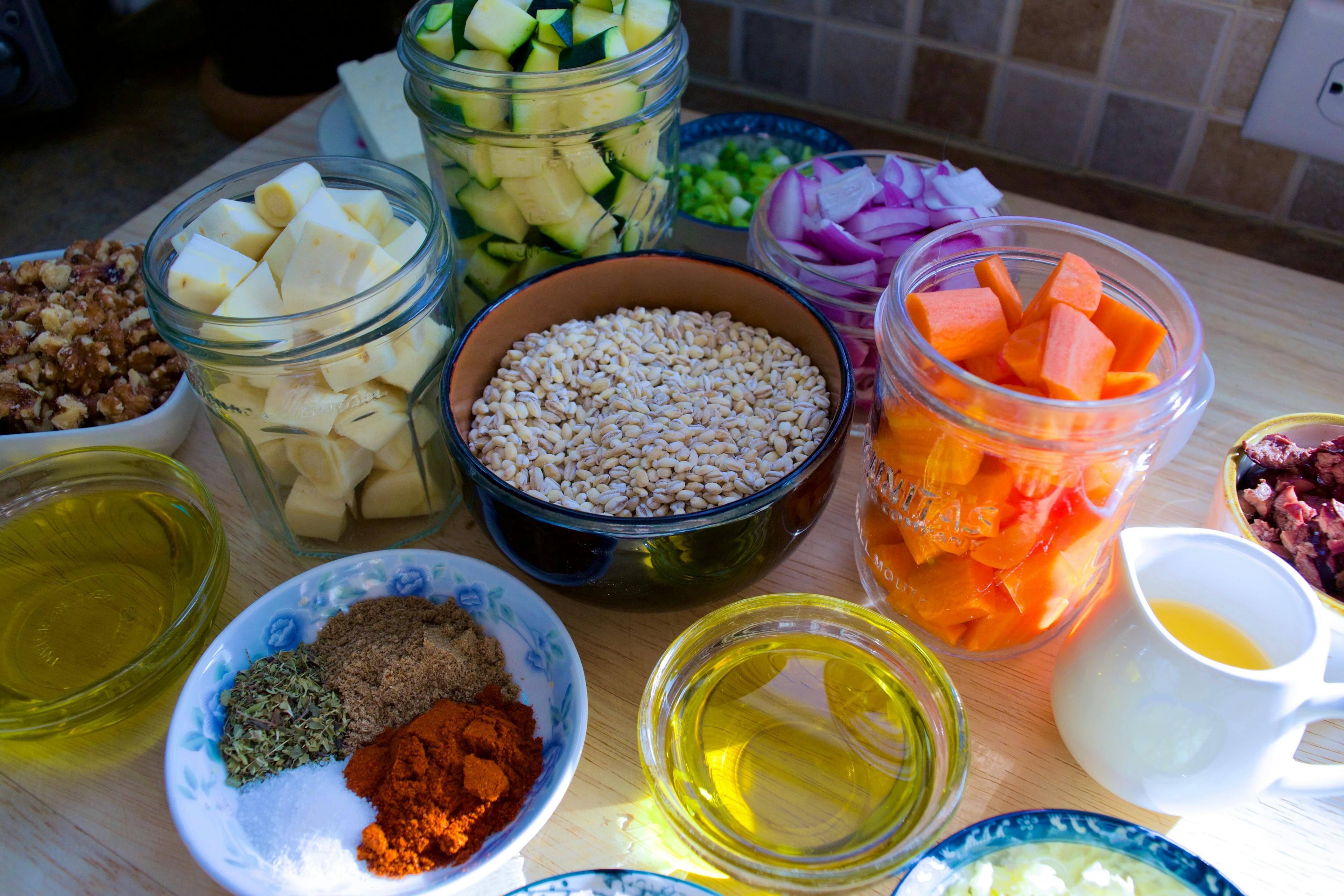 Ingredients in jars and dishes- zucchini, parsnips, red onion, walnuts, dried barley, spices, carrots, olive oil, and olives are all visible.