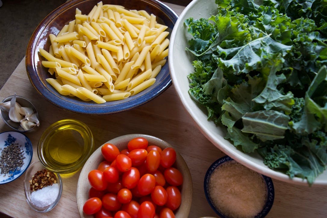 Ingredients in bowls - pasta, kale, cherry tomatoes, olive oil, cheese, garlic, spices.