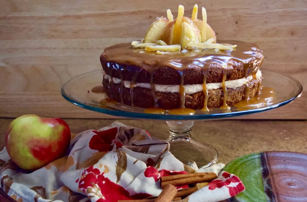 The cake on a clear glass cake stand. There is a napkin with an apple and some cinnamon sticks in the foreground. 